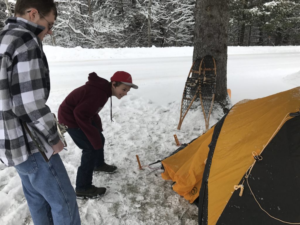 Two students peer into a tent  set up in a snowy landscape
