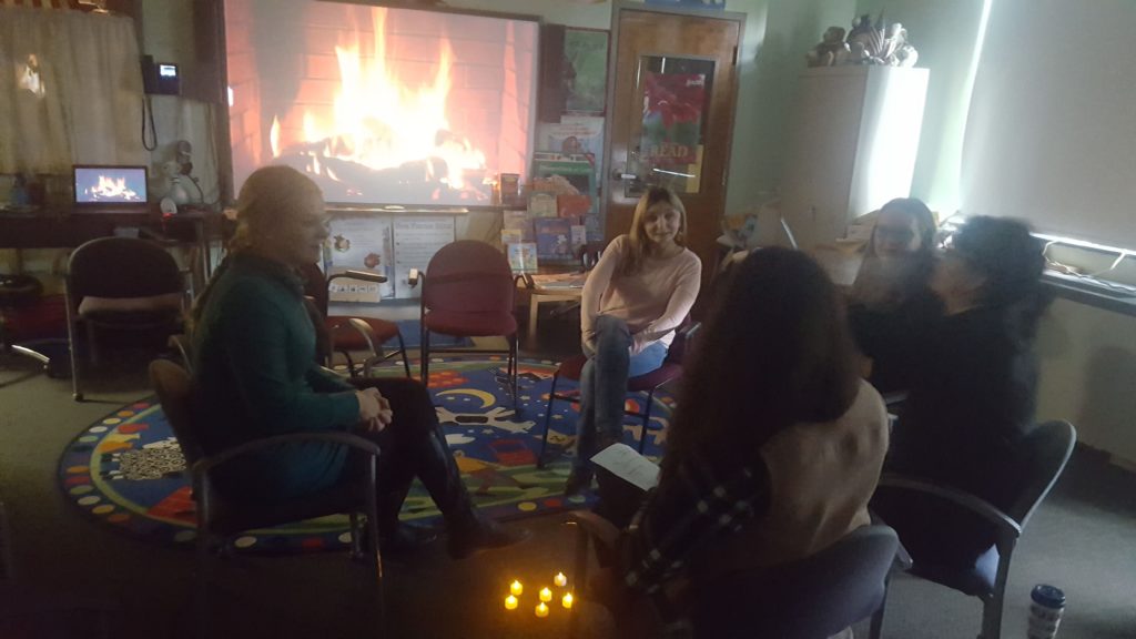 A group of women sit chatting in a circle of chairs in a darkened classroom. Behind them, a video of a roaring fireplace is projected onto a whiteboard from a laptop.