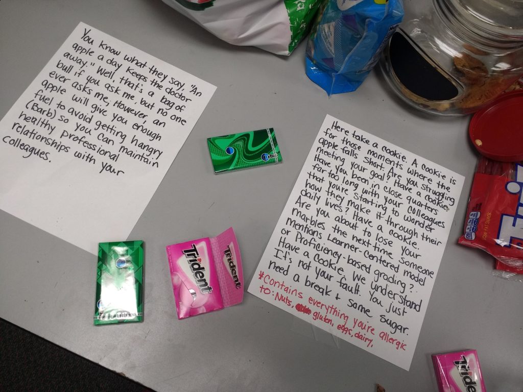 Left sheet of paper: "You know what they say, 'An apple a day keeps the doctor away.' Well, that's a bag of bull if you ask me, but no one ever asks me. However, an apple will give you enough fuel to avoid getting hangry (Barb) so you can maintain healthy professional relationships with your colleagues." Righthand sheet of paper: "Here take a cookie. A cookie is for those moments where the apple falls short. Are you struggling meeting your goal? Have a cookie. Have you been in close quarters for too long with your colleagues that you're starting to wonder how they make it through their daily lives? Have a cookie. Are you about to lose your marbles the next time someone mentions Learn-Centered Model or Proficiency-Based grading? Have a cookie. We understand it's not your fault. You just need a break and some sugar. *Contains everything you're allergic to: nuts, gluten, eggs, dairy."
