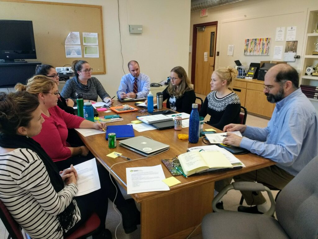 Teachers sit around a table covered with papers and computers talking.