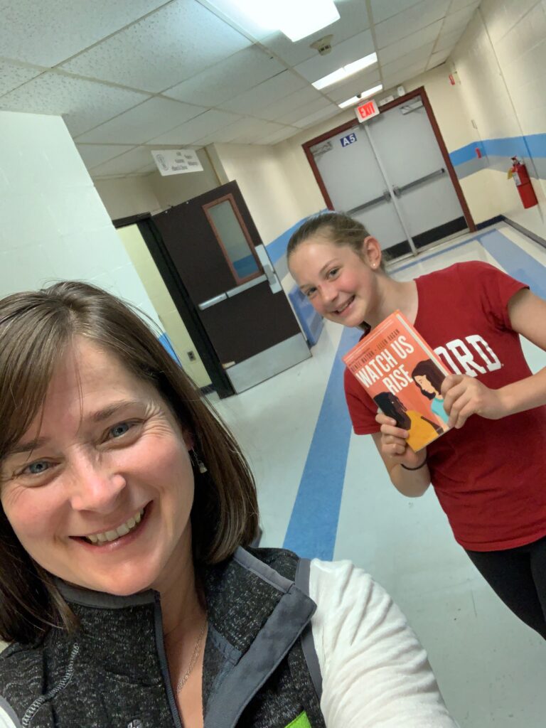 Jeanie Phillips, a whitewoman with straight brown hair, takes a selfie with Abby Bunting, a white 9th grader standing behind her, grinning and holding up a copy of Watch Us Rise