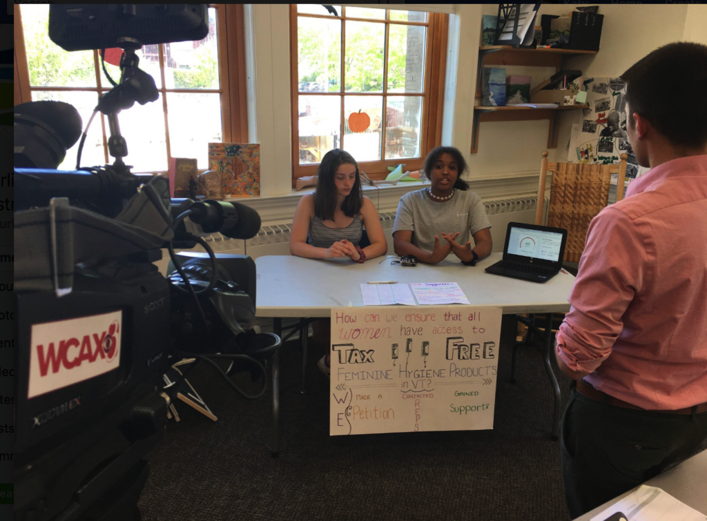 Two students sit behind a desk in a well-lit classroom, speaking to an adult in front of them. A WCAX news camera is also trained on them. From a critical lens on project-based learning.