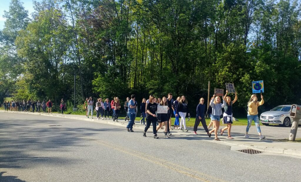 Large group of students walk on sidewalk holding signs. Confronting climate change