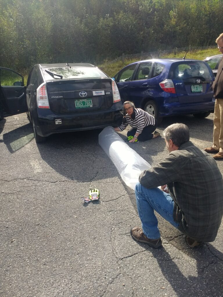 2 adults hold a plastic tube-shaped bag to a car's exhaust pipe, confronting climate change