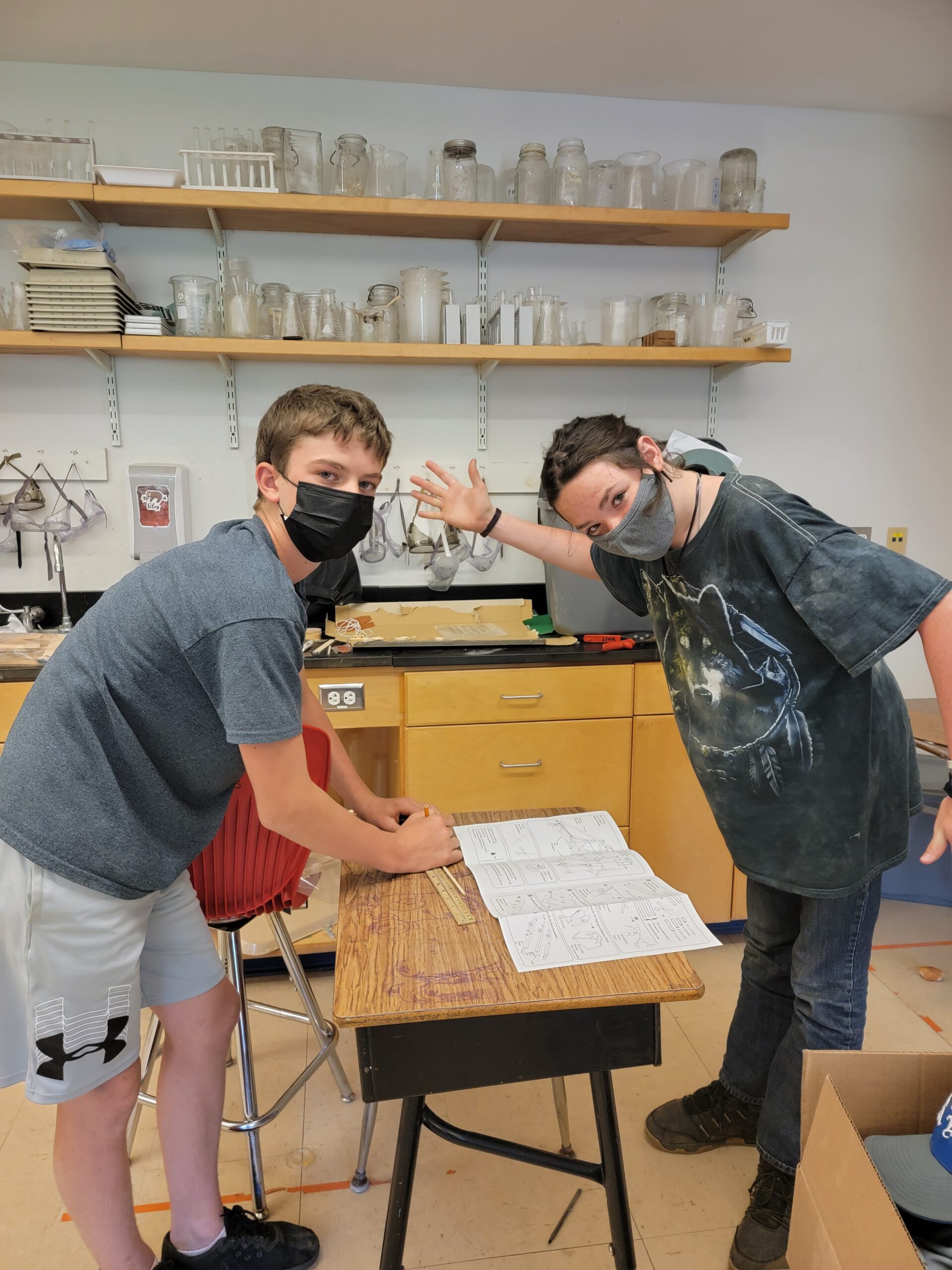 Two masked students pause their science work to wave at the camera