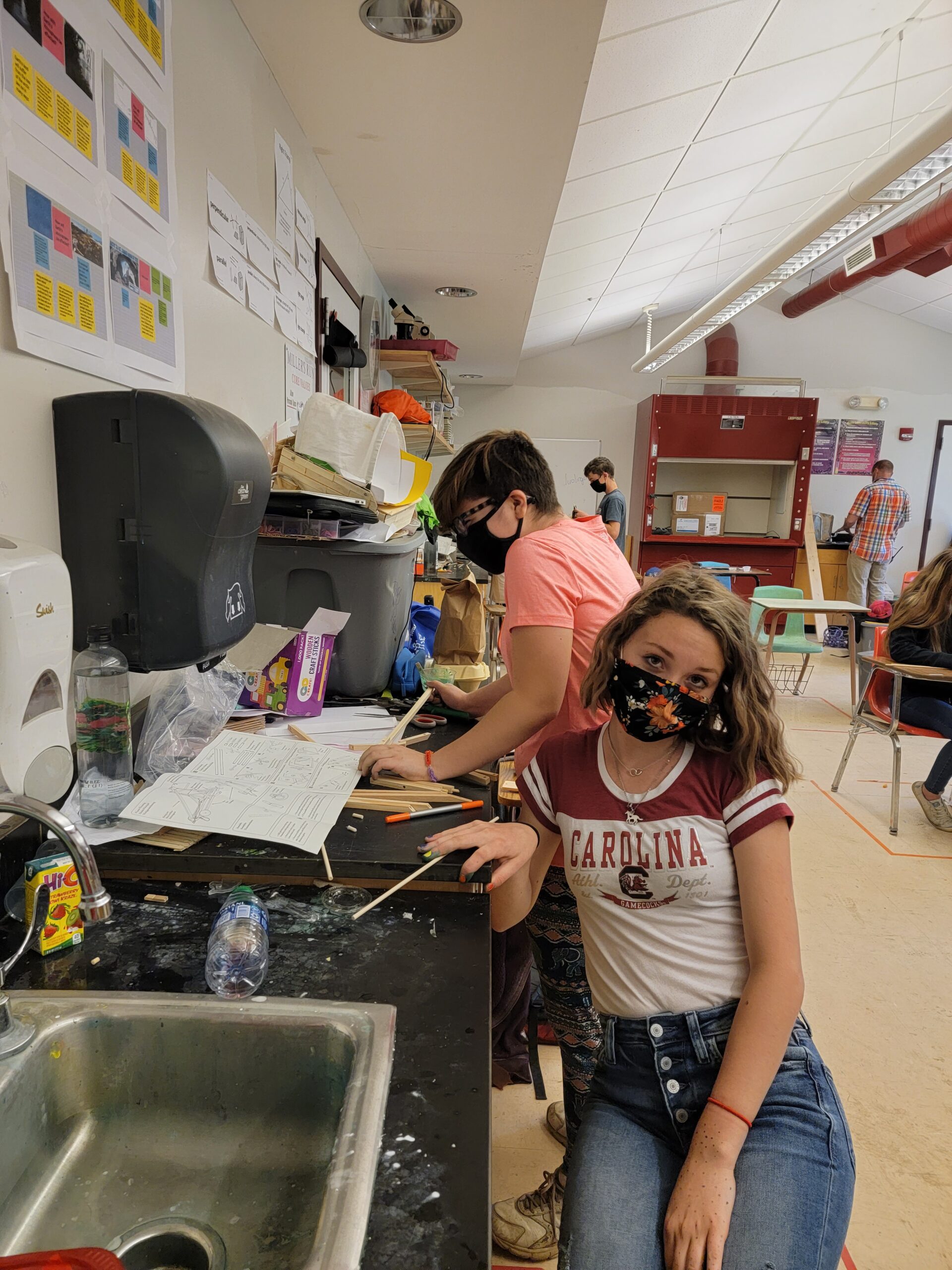 One masked student works on science while another poses for the camera