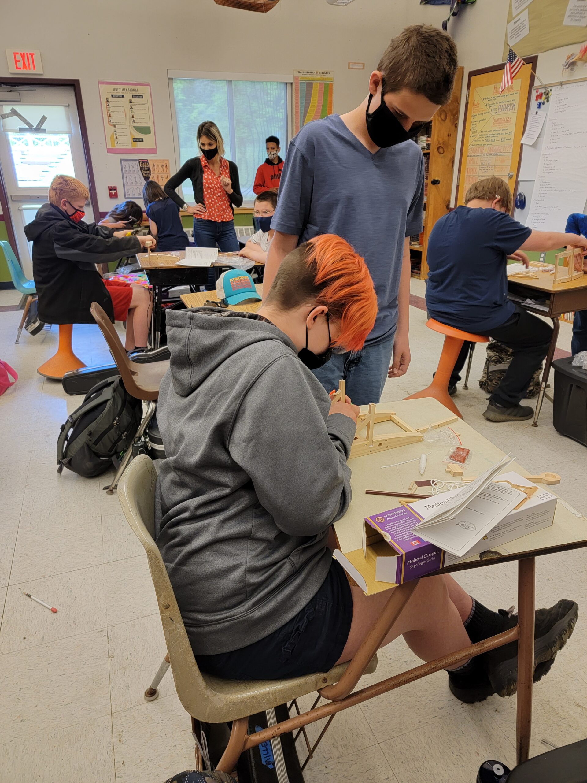 A student with orange-pink hair works at their desk while another looks on