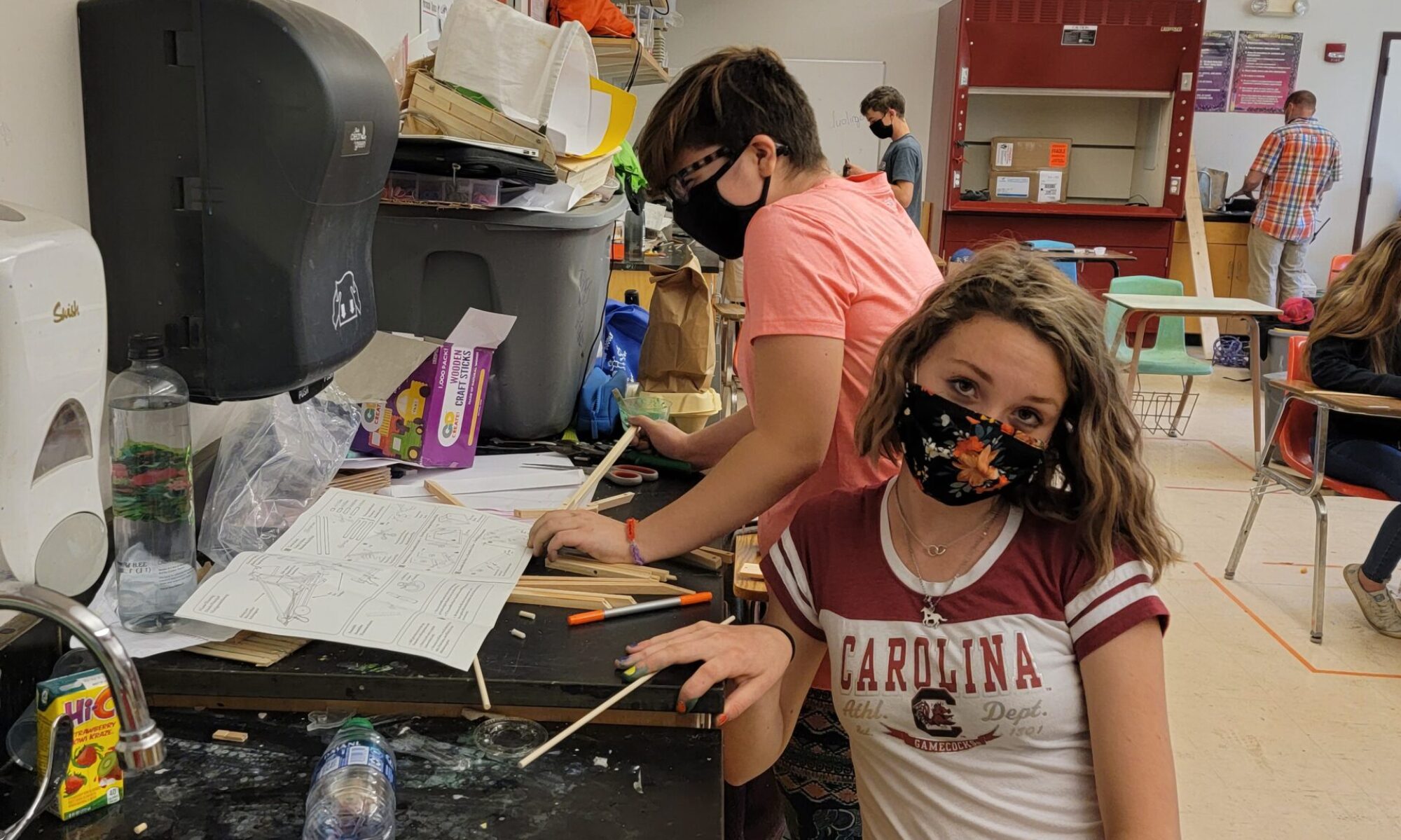 One masked student works on science while another poses for the camera