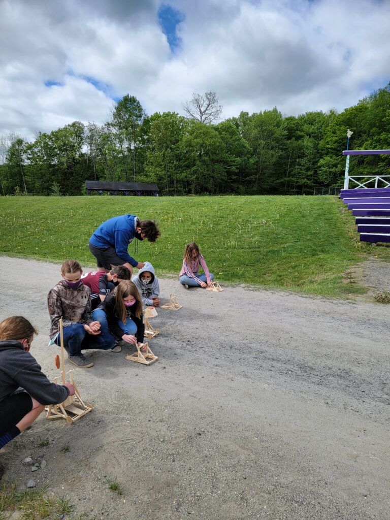 Students crouch in the dirt near tiny wooden catapults