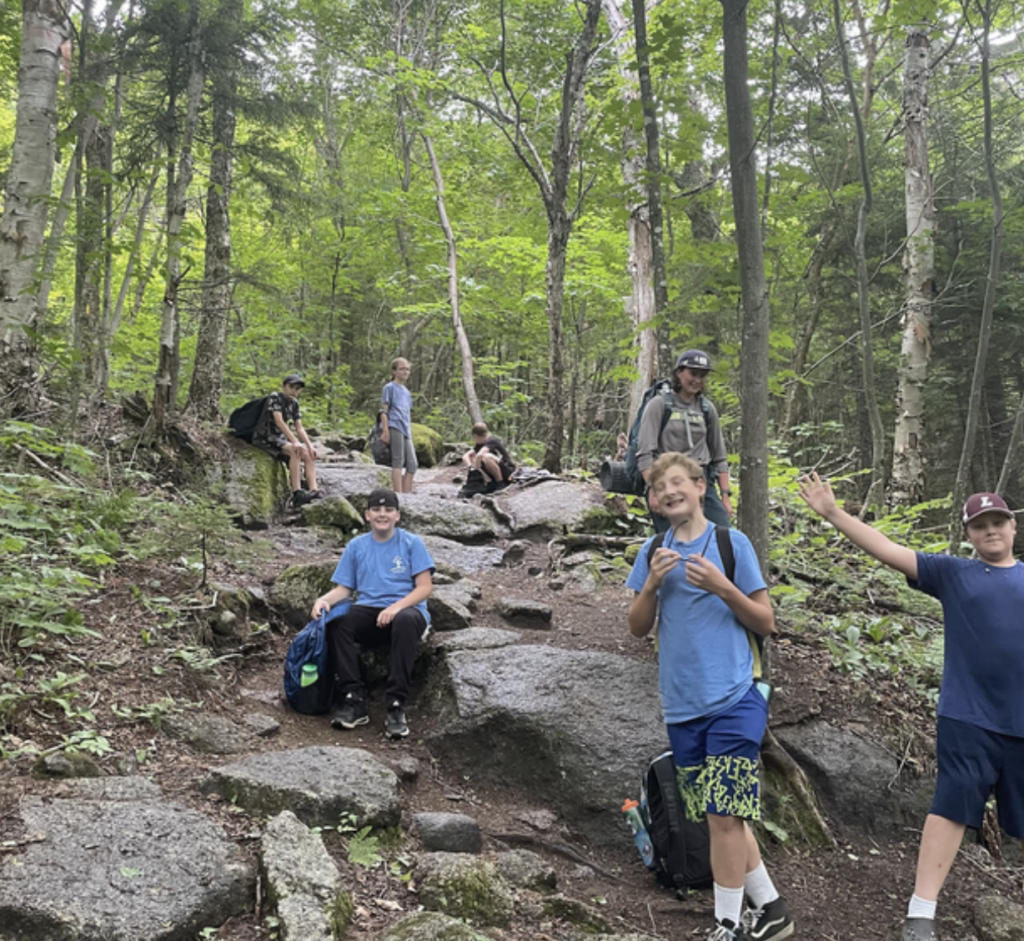 Students rest on a rocky path through the woods. One salutes the camera with open arms.