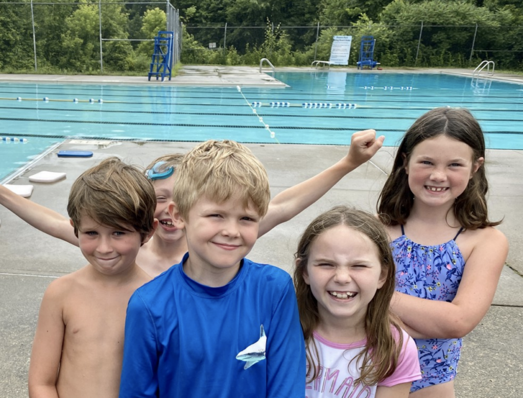 A group of children in swimwear stand next to a still community pool, smiling at the camera.