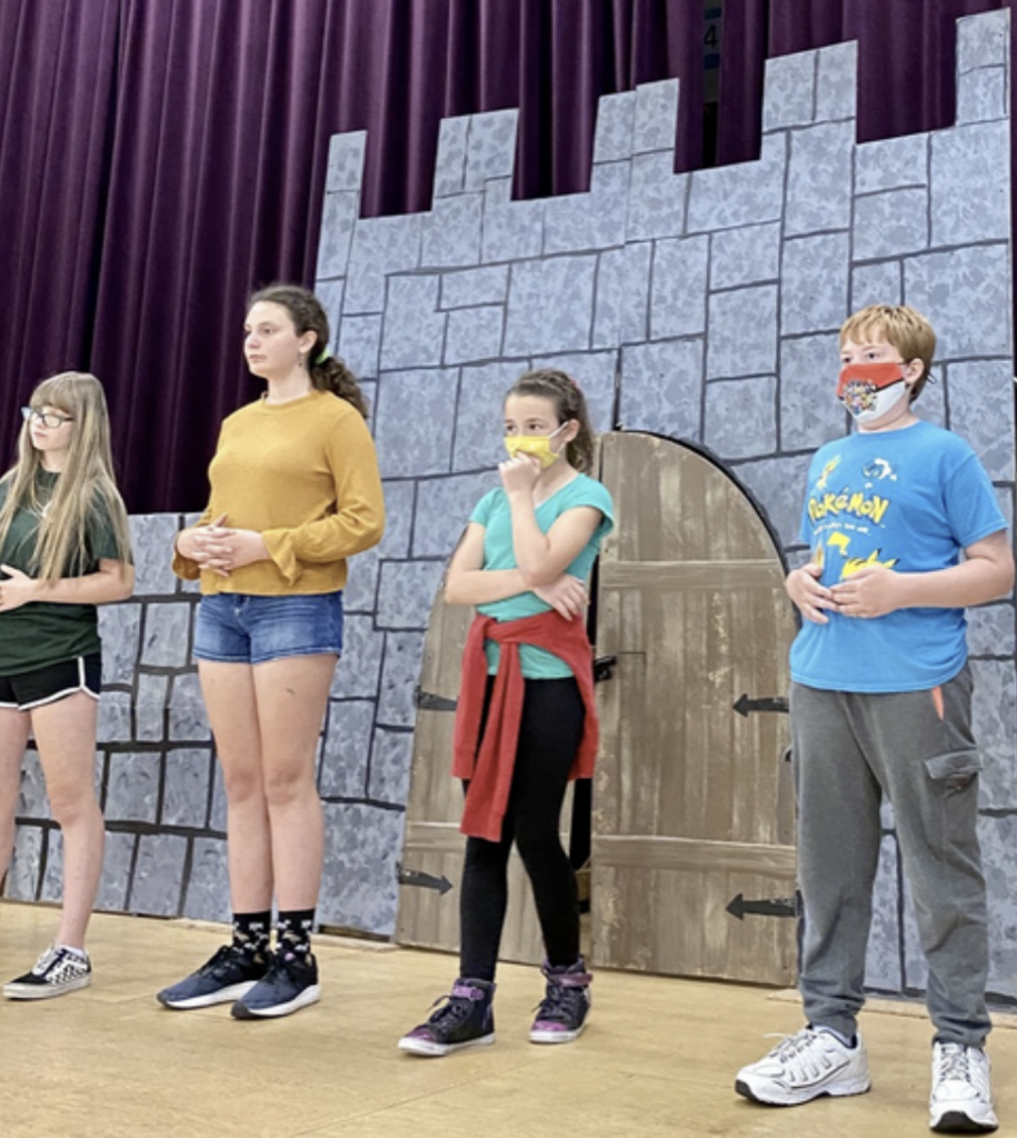 Four children stand on a school stage, looking off camera, in front of a castle backdrop. Two wear cloth masks.