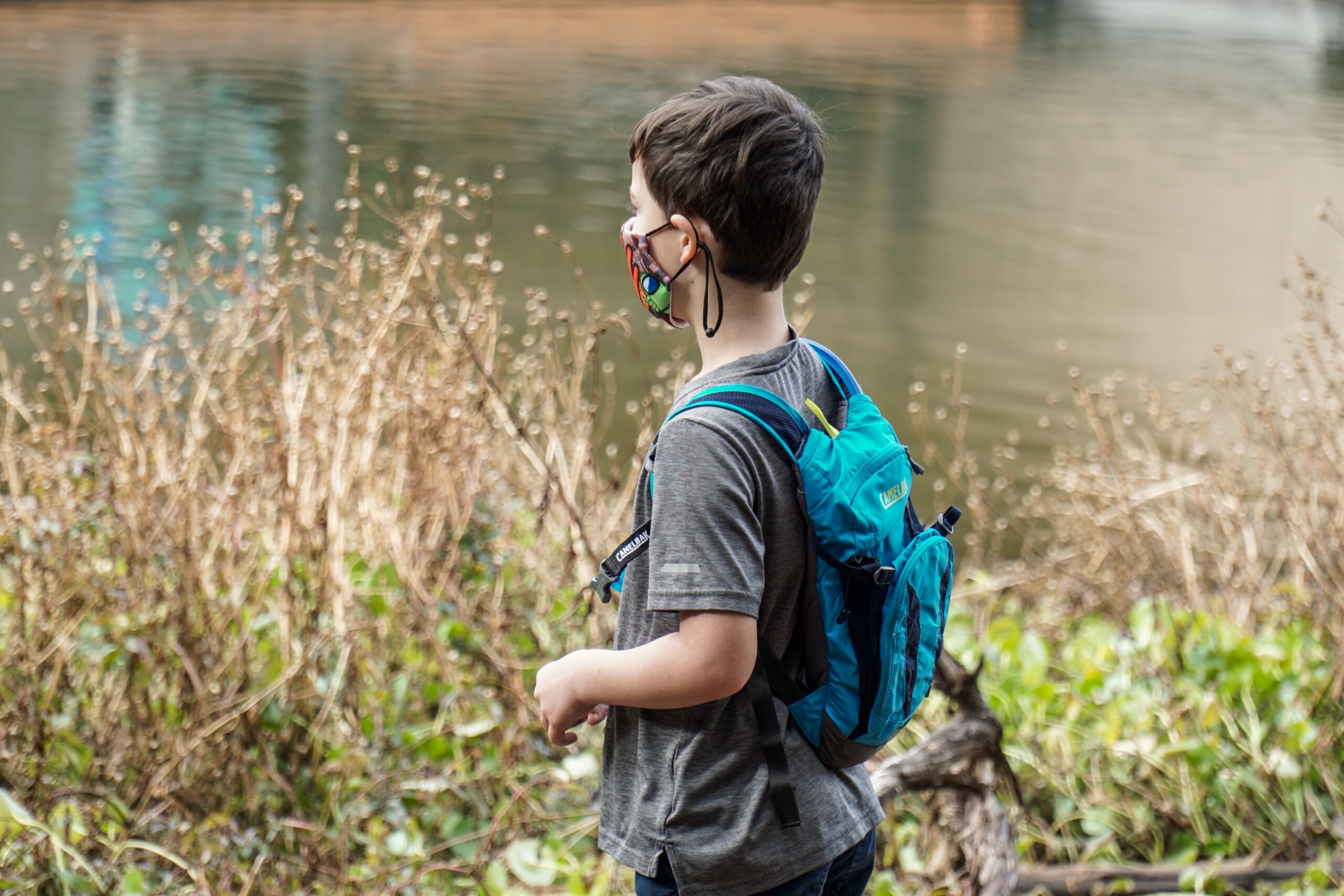 A student in a cloth mask, wearing a backpack, walks along the edge of a pond.