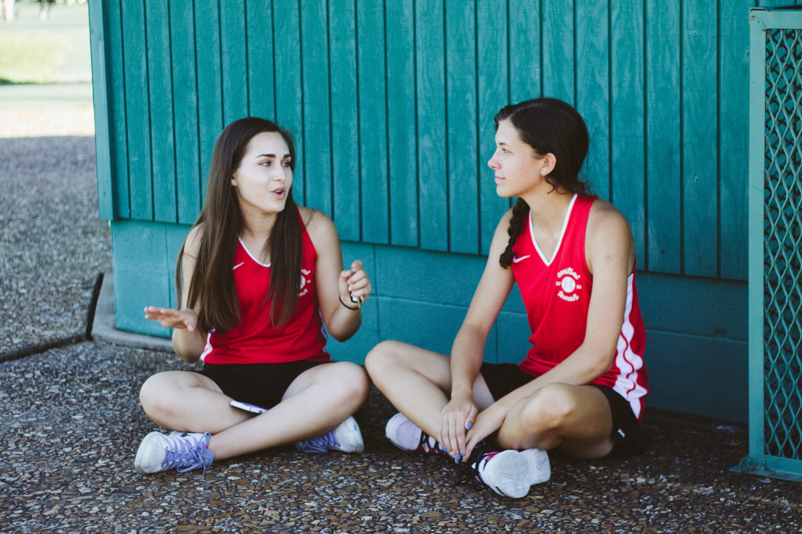 Two students in P.E. gear sit cross-legged next to a teal wall, in the shade.