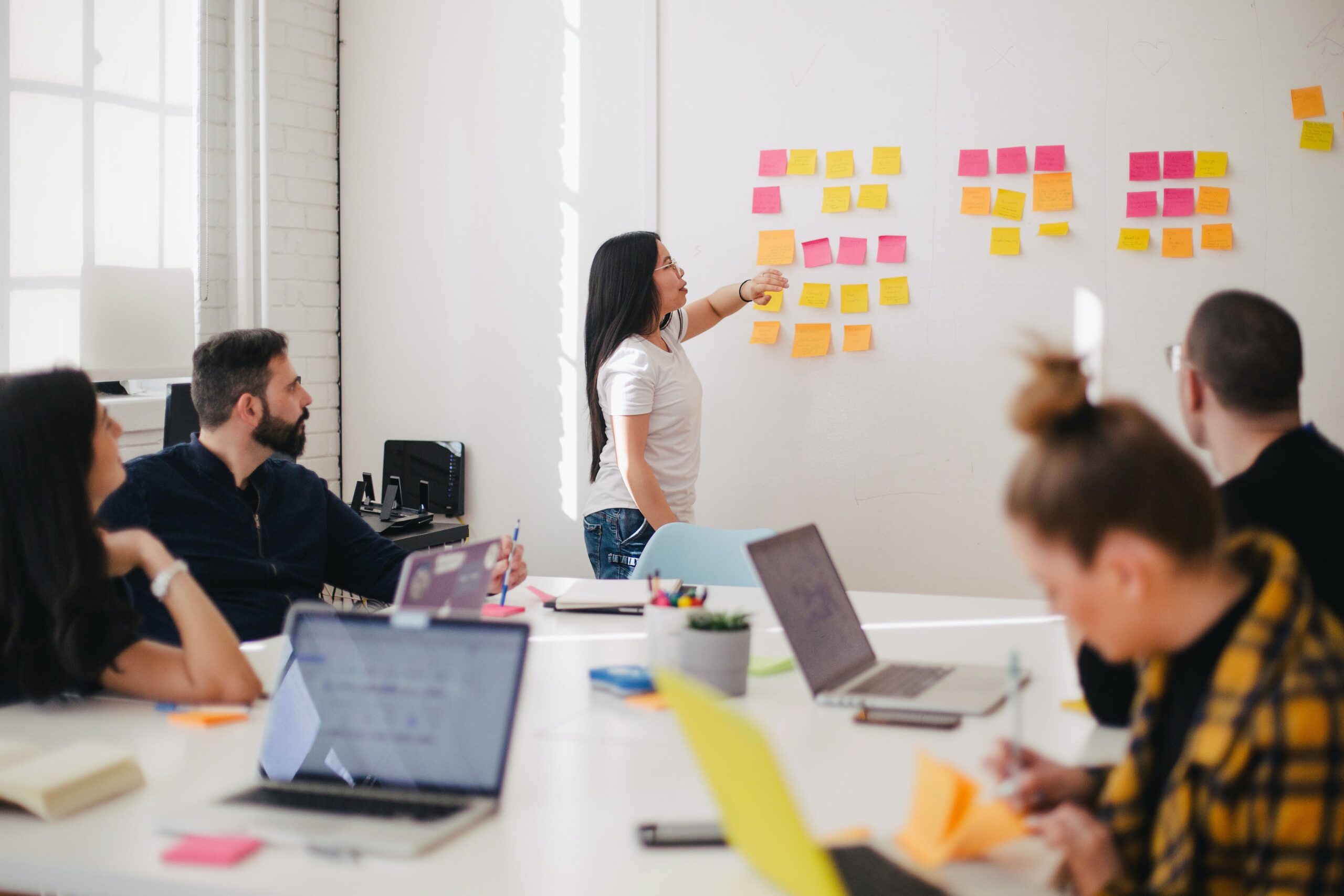 A group of adults gather around a conference room table as a woman with long straight black hair gestures to a grid of multi-colored post-it notes on the wall at the front of the room