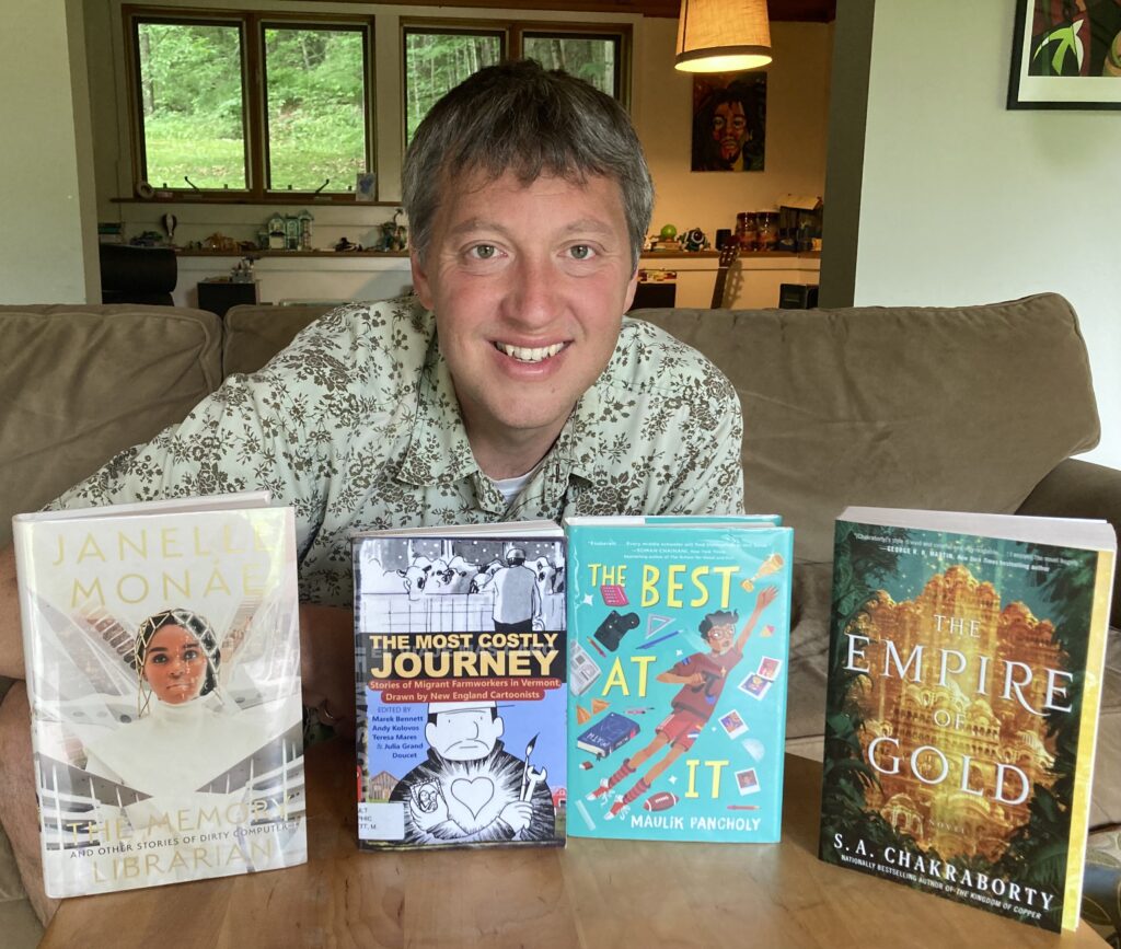 A man with salt-and-pepper short hair poses with four books on a coffee table