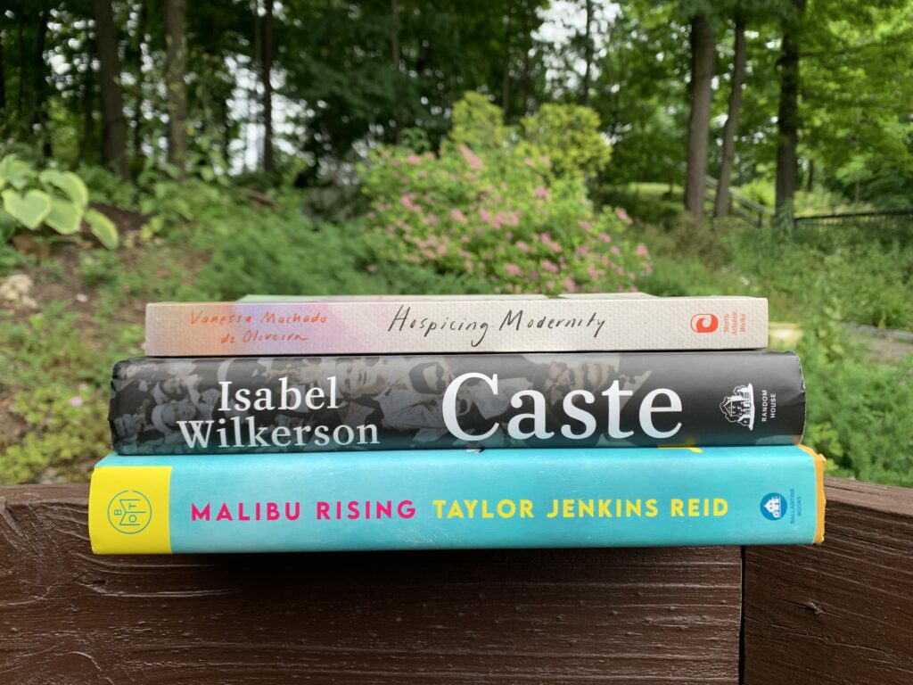 a picture of three books outside in a garden: Malibu Rising, Caste, and Hospicing Modernity.