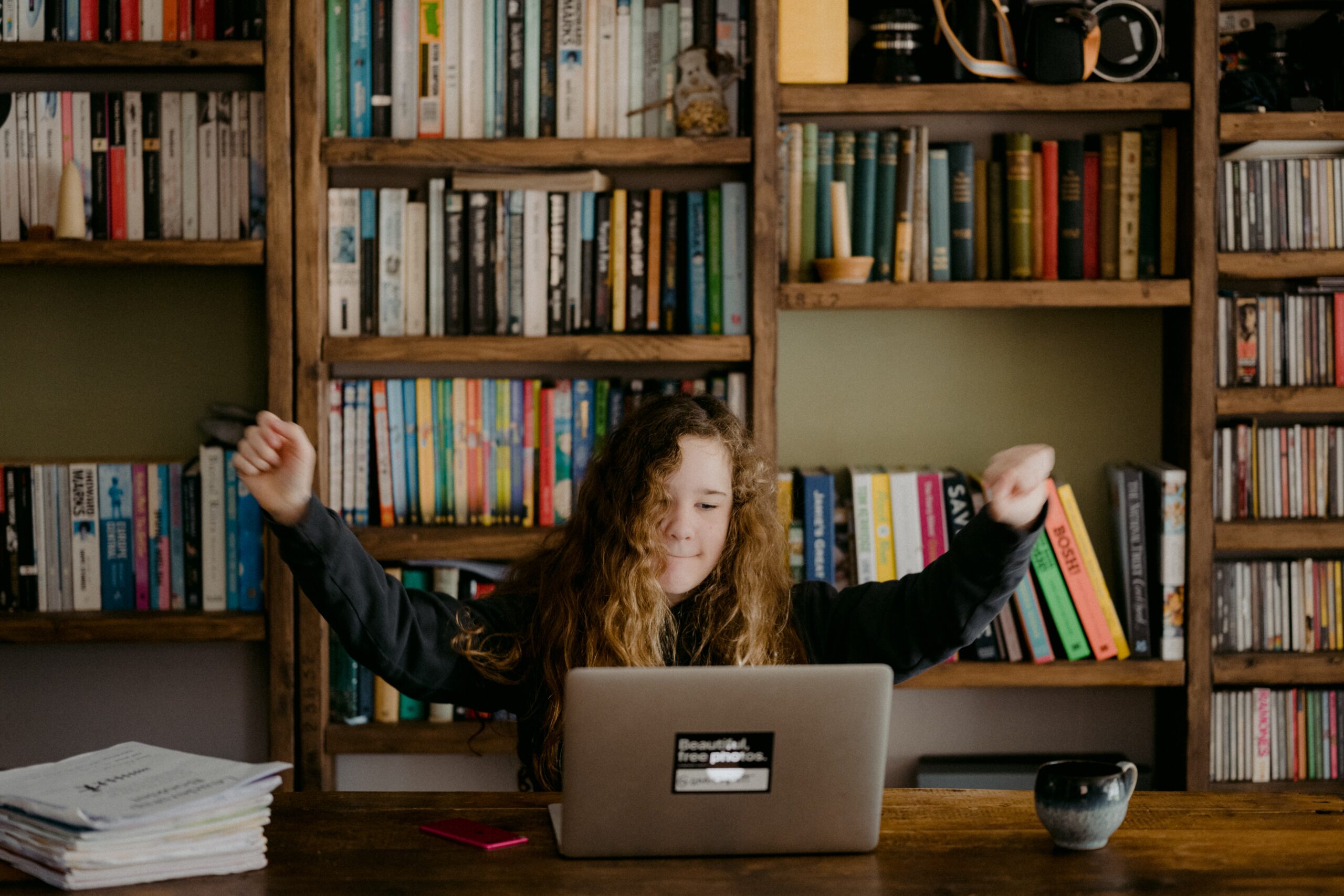 A student in a hoodie sits at a laptop and raises their arms in triumph