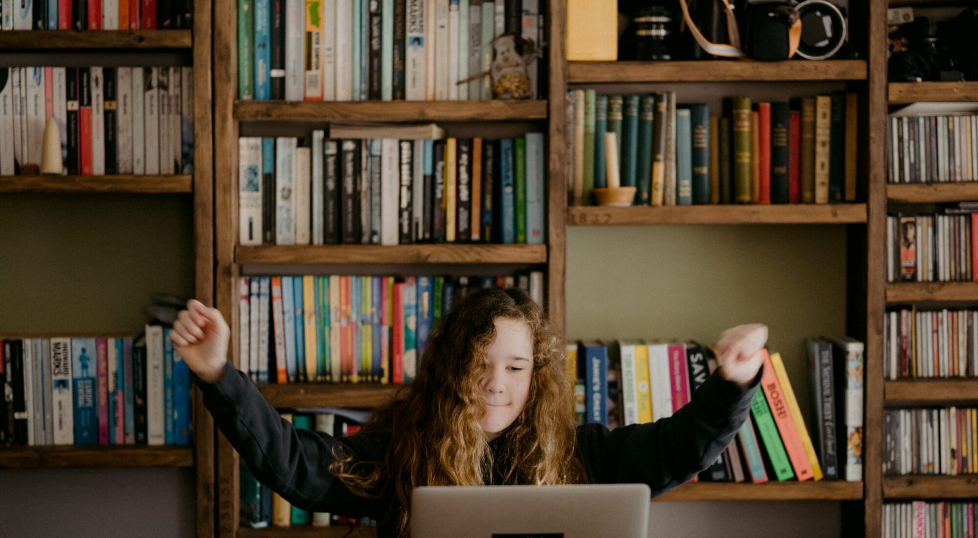 A student in a hoodie sits at a laptop and raises their arms in triumph