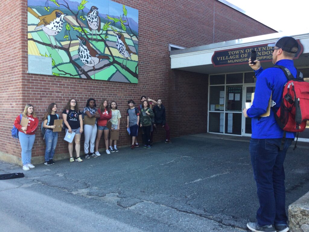 A group of students stand along the wall of another building, under a mural