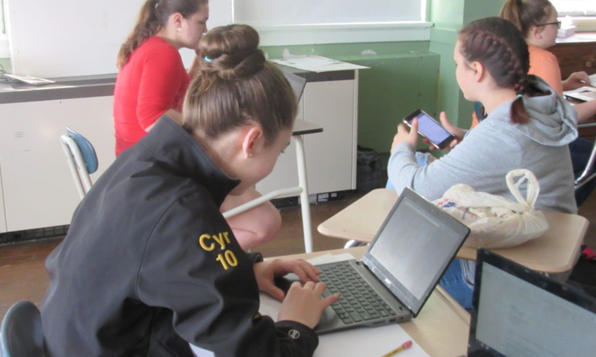 One student works at a laptop while two others work on a phone